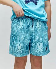 A person wears Psycho Bunny KIDS CHUCK ALL OVER PRINT LIGHTWEIGHT SWIM TRUNK (KB1000002), featuring a wavy dark blue and white bunny print, quick-dry fabric, and a drawstring waistband, paired with a matching blue t-shirt.