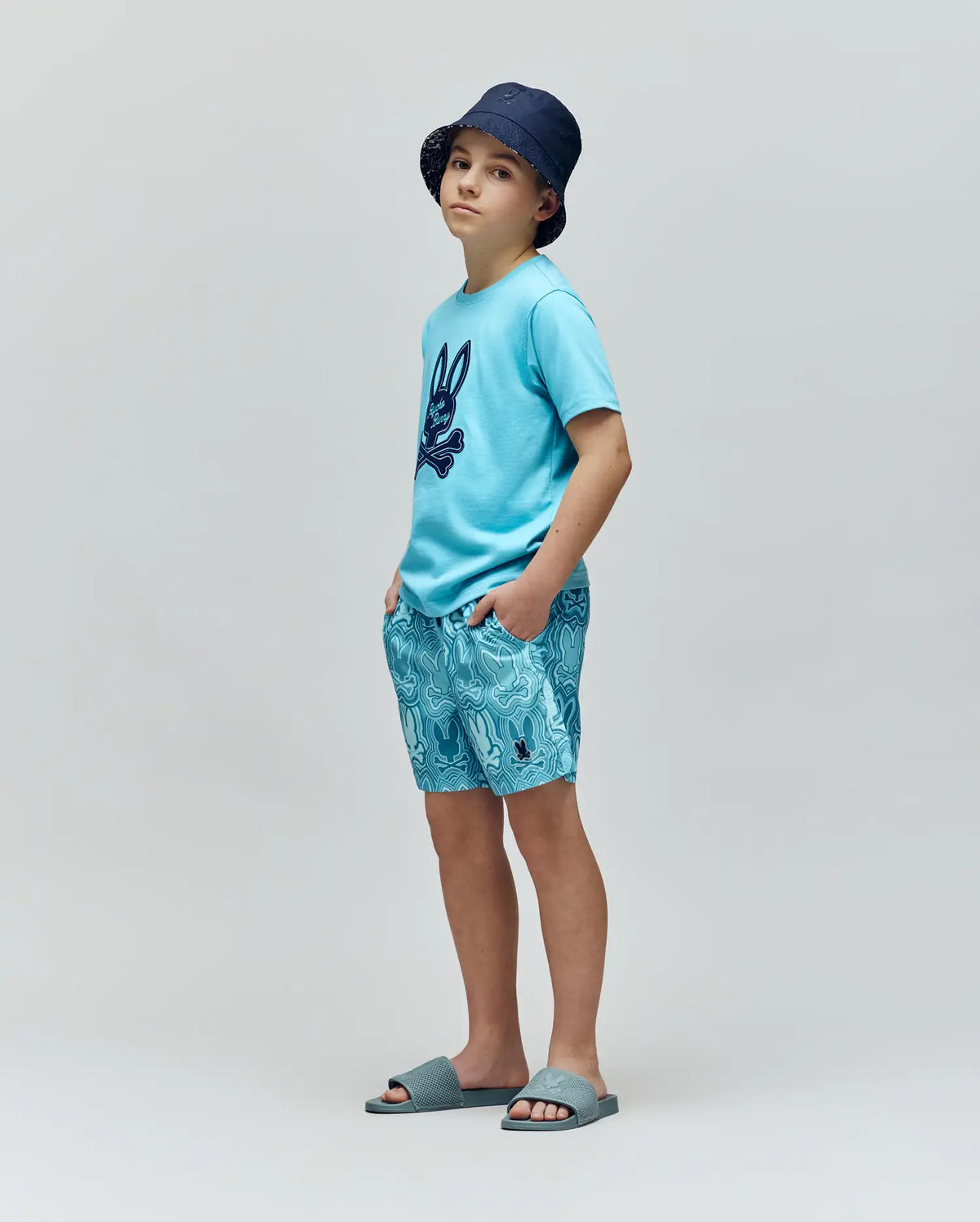 A young boy wearing a navy bucket hat, light blue Psycho Bunny T-shirt with a dark bunny print, KIDS CHUCK ALL OVER PRINT LIGHTWEIGHT SWIM TRUNK by Psycho Bunny, and blue slide sandals stands against a plain gray background.