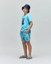 A young boy wearing a navy bucket hat, light blue Psycho Bunny T-shirt with a dark bunny print, KIDS CHUCK ALL OVER PRINT LIGHTWEIGHT SWIM TRUNK by Psycho Bunny, and blue slide sandals stands against a plain gray background.