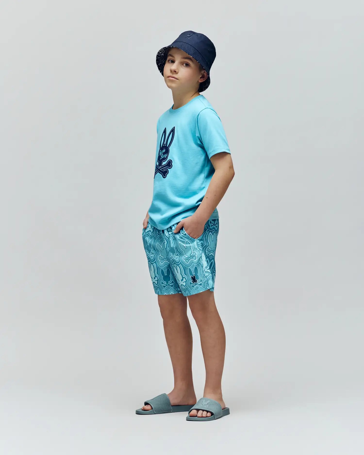 A young boy wearing a navy bucket hat, light blue Psycho Bunny T-shirt with a dark bunny print, KIDS CHUCK ALL OVER PRINT LIGHTWEIGHT SWIM TRUNK by Psycho Bunny, and blue slide sandals stands against a plain gray background.