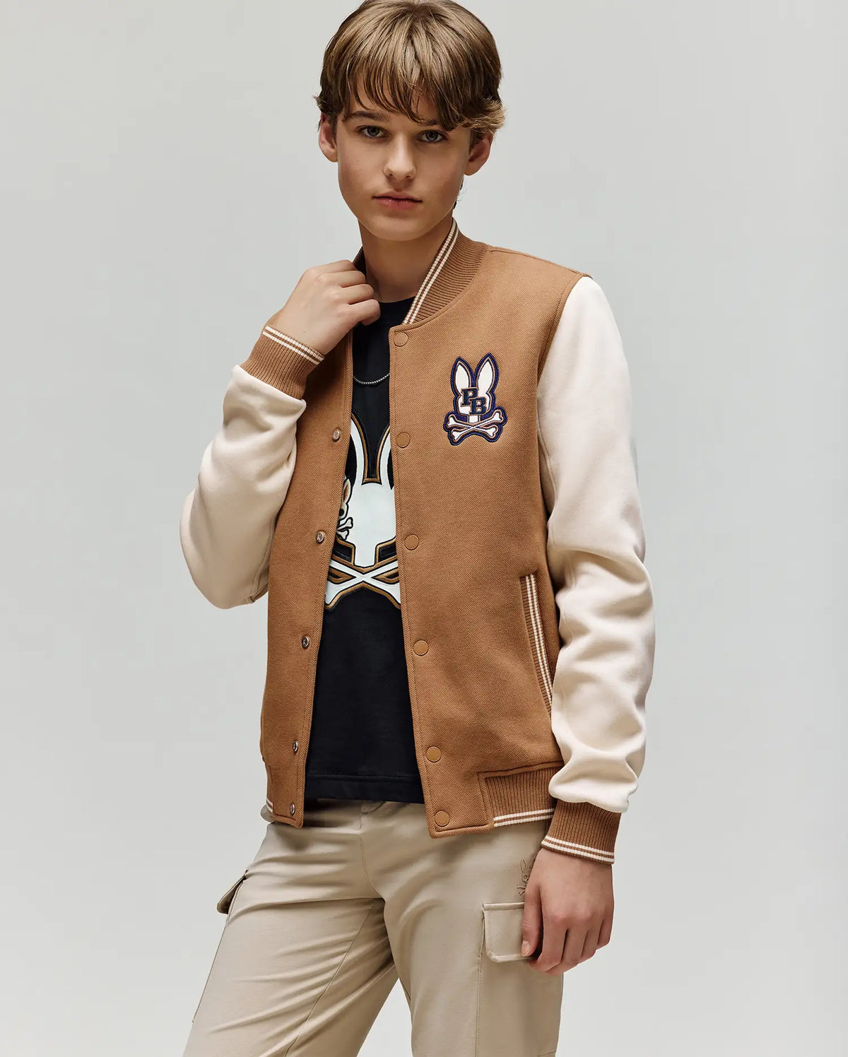 A young person wears the Psycho Bunny KIDS WADE KNIT BOMBER (KO1100001) in brown and beige, featuring a bunny patch, with a matching black bunny graphic shirt and beige pants, standing against a light gray background.