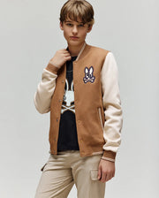 A young person wears the Psycho Bunny KIDS WADE KNIT BOMBER (KO1100001) in brown and beige, featuring a bunny patch, with a matching black bunny graphic shirt and beige pants, standing against a light gray background.