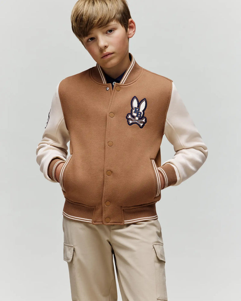 A young boy with light brown hair models the Psycho Bunny KIDS WADE KNIT BOMBER (KO1100001), a varsity-style jacket with a rabbit patch, paired with beige cargo pants, standing hands-in-pockets against a plain background.
