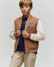 A young boy wears the Psycho Bunny KIDS WADE KNIT BOMBER (KO1100001) in tan and cream with a bunny patch, paired with a navy polo shirt and beige pants, standing against a plain background and looking at the camera neutrally.