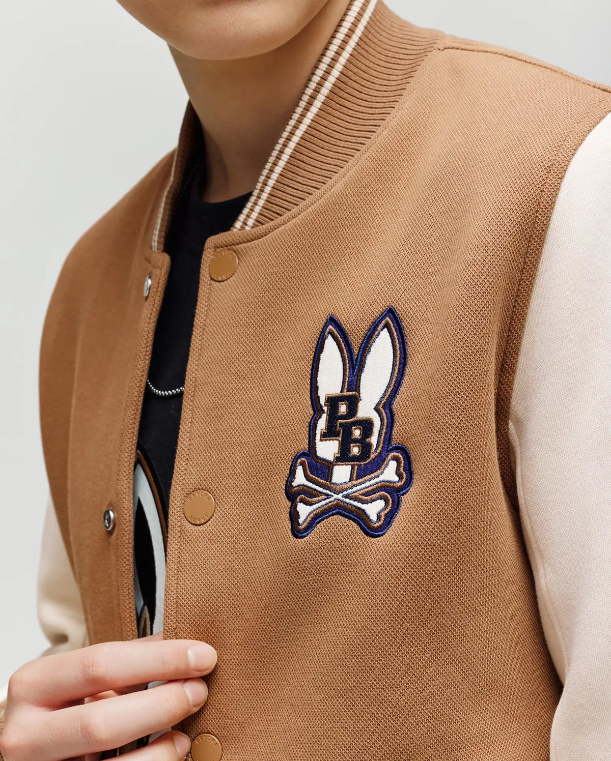 A child wears the Psycho Bunny KIDS WADE KNIT BOMBER (KO1100001) in tan and cream, featuring a bunny head and crossbones patch above "PB" on the chest. The child's face is partially out of frame.