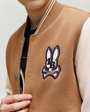 A child wears the Psycho Bunny KIDS WADE KNIT BOMBER (KO1100001) in tan and cream, featuring a bunny head and crossbones patch above "PB" on the chest. The child's face is partially out of frame.
