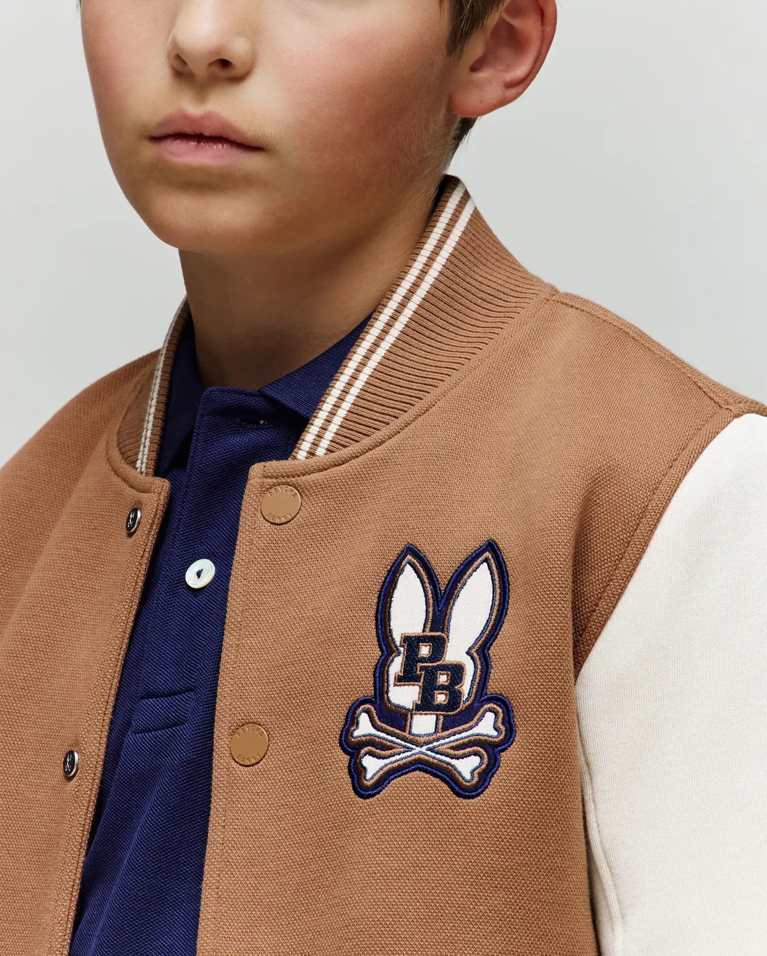 A young boy wears the Psycho Bunny KIDS WADE KNIT BOMBER (KO1100001) in tan and white, featuring a blue PB bunny and crossbones patch over a blue collared shirt. Only the lower half of his face is visible.