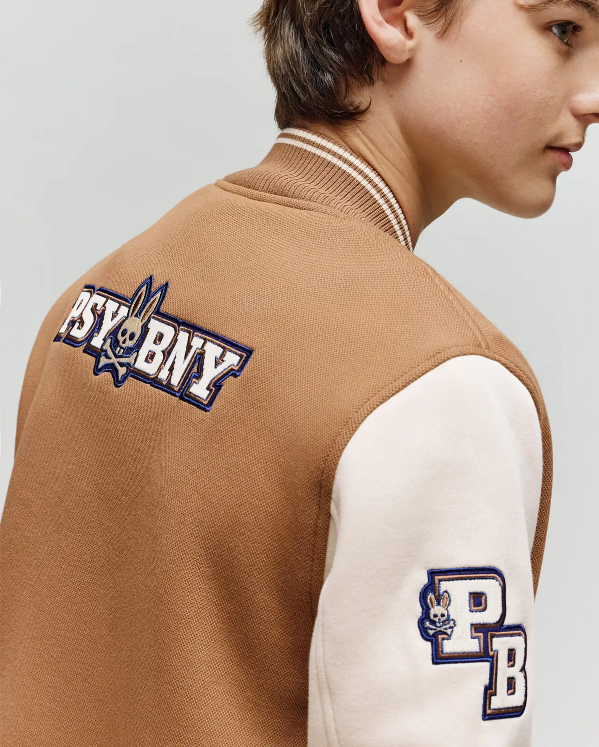 A young person models the Psycho Bunny KIDS WADE KNIT BOMBER (KO1100001), a premium brown and cream varsity jacket featuring "PSYBNY" and bunny logo on the back, plus a "PB" sleeve patch. Shown from the side, slightly turned away.
