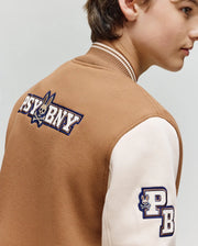 A young person models the Psycho Bunny KIDS WADE KNIT BOMBER (KO1100001), a premium brown and cream varsity jacket featuring "PSYBNY" and bunny logo on the back, plus a "PB" sleeve patch. Shown from the side, slightly turned away.