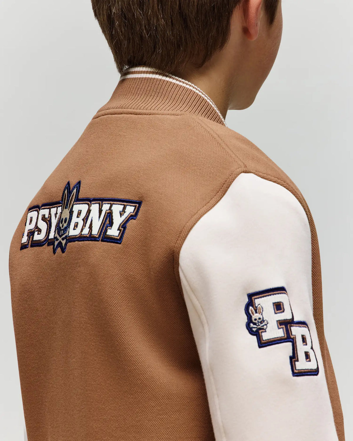 A child wears the Psycho Bunny KIDS WADE KNIT BOMBER (KO1100001) in brown and white, featuring "PSYBNY" and a bunny logo on the back, plus a "PB" bunny patch on the sleeve. Shown from behind and slightly to the side. Premium cotton blend.