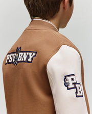 A child wears the Psycho Bunny KIDS WADE KNIT BOMBER (KO1100001) in brown and white, featuring "PSYBNY" and a bunny logo on the back, plus a "PB" bunny patch on the sleeve. Shown from behind and slightly to the side. Premium cotton blend.