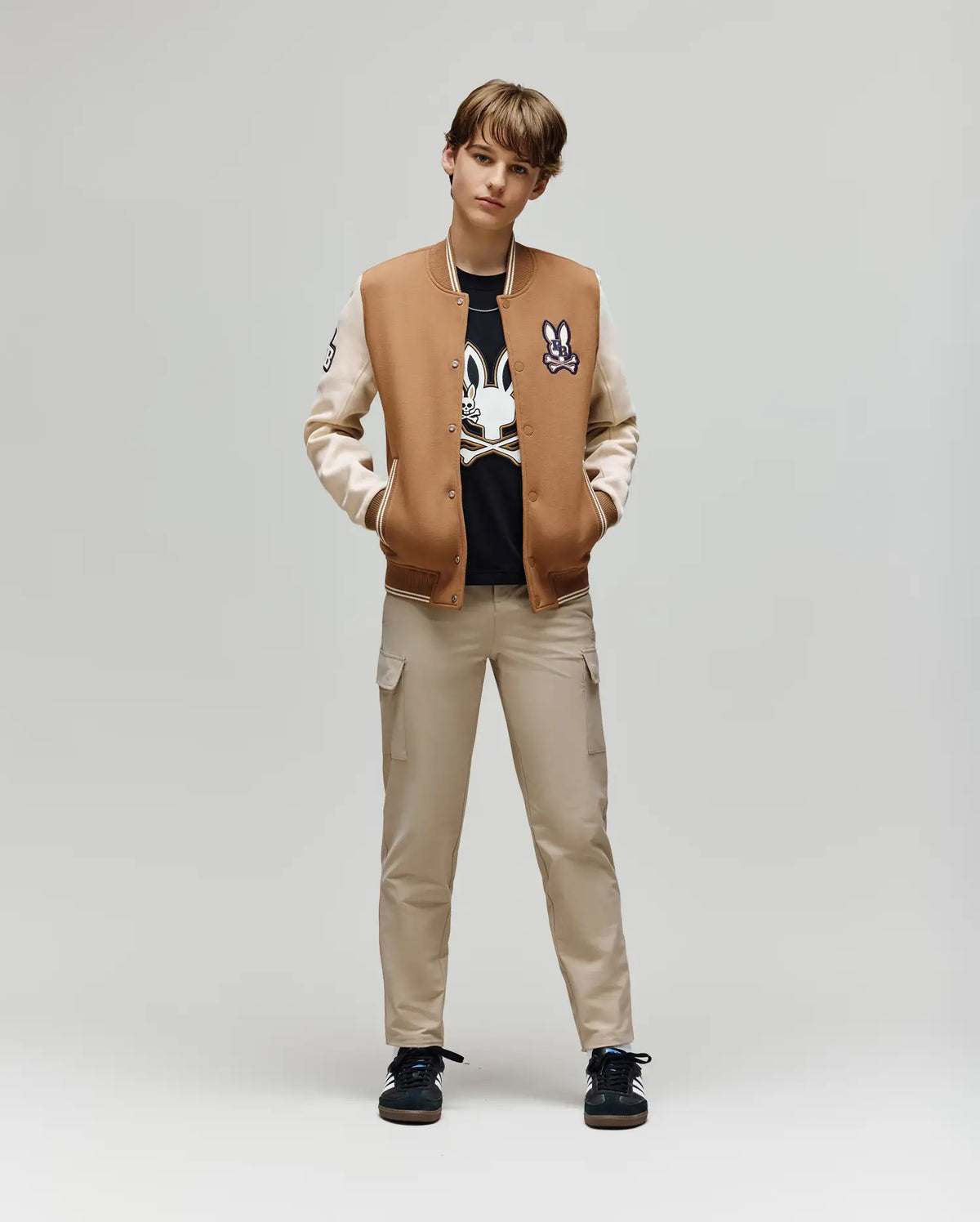 A young person stands facing forward, hands in pockets, wearing the Psycho Bunny KIDS WADE KNIT BOMBER (KO1100001) over a black graphic tee, beige cargo pants, and black sneakers against a plain light background.