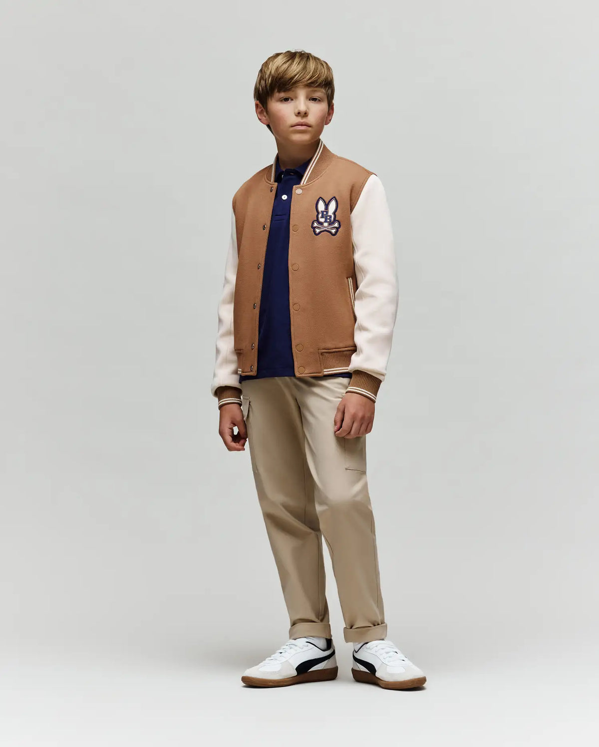 A young boy wears the Psycho Bunny KIDS WADE KNIT BOMBER (KO1100001), a premium cotton-blend tan and white varsity jacket with a cartoon patch, styled with a navy shirt, beige pants, and white sneakers. He stands calmly against a plain background.