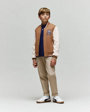 A young boy wears the Psycho Bunny KIDS WADE KNIT BOMBER (KO1100001), a premium cotton-blend tan and white varsity jacket with a cartoon patch, styled with a navy shirt, beige pants, and white sneakers. He stands calmly against a plain background.