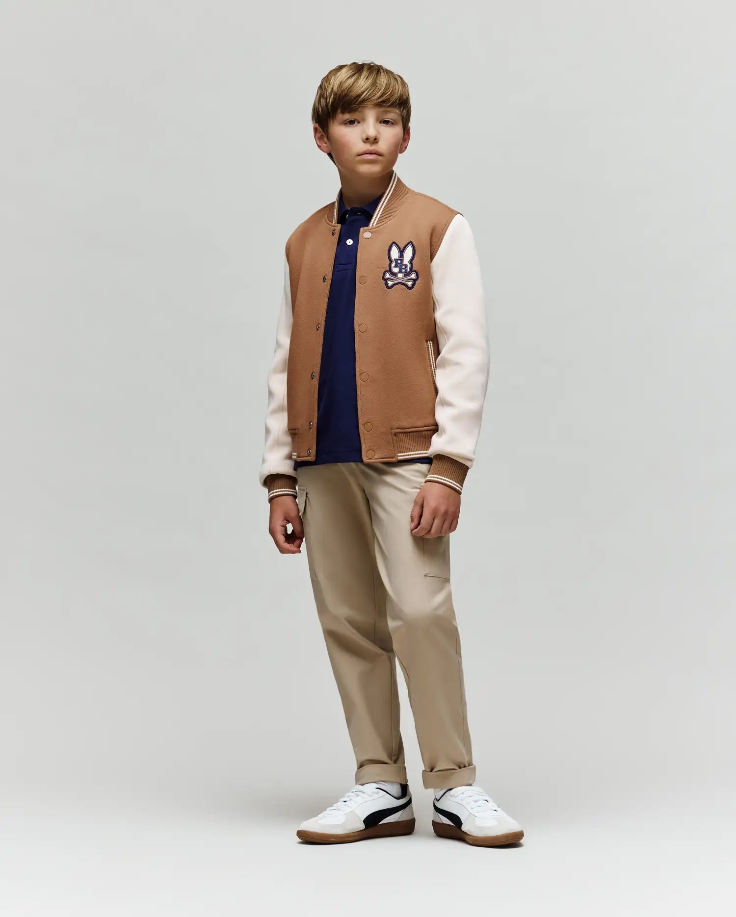 A young boy wears the Psycho Bunny KIDS WADE KNIT BOMBER (KO1100001), a premium cotton-blend tan and white varsity jacket with a cartoon patch, styled with a navy shirt, beige pants, and white sneakers. He stands calmly against a plain background.