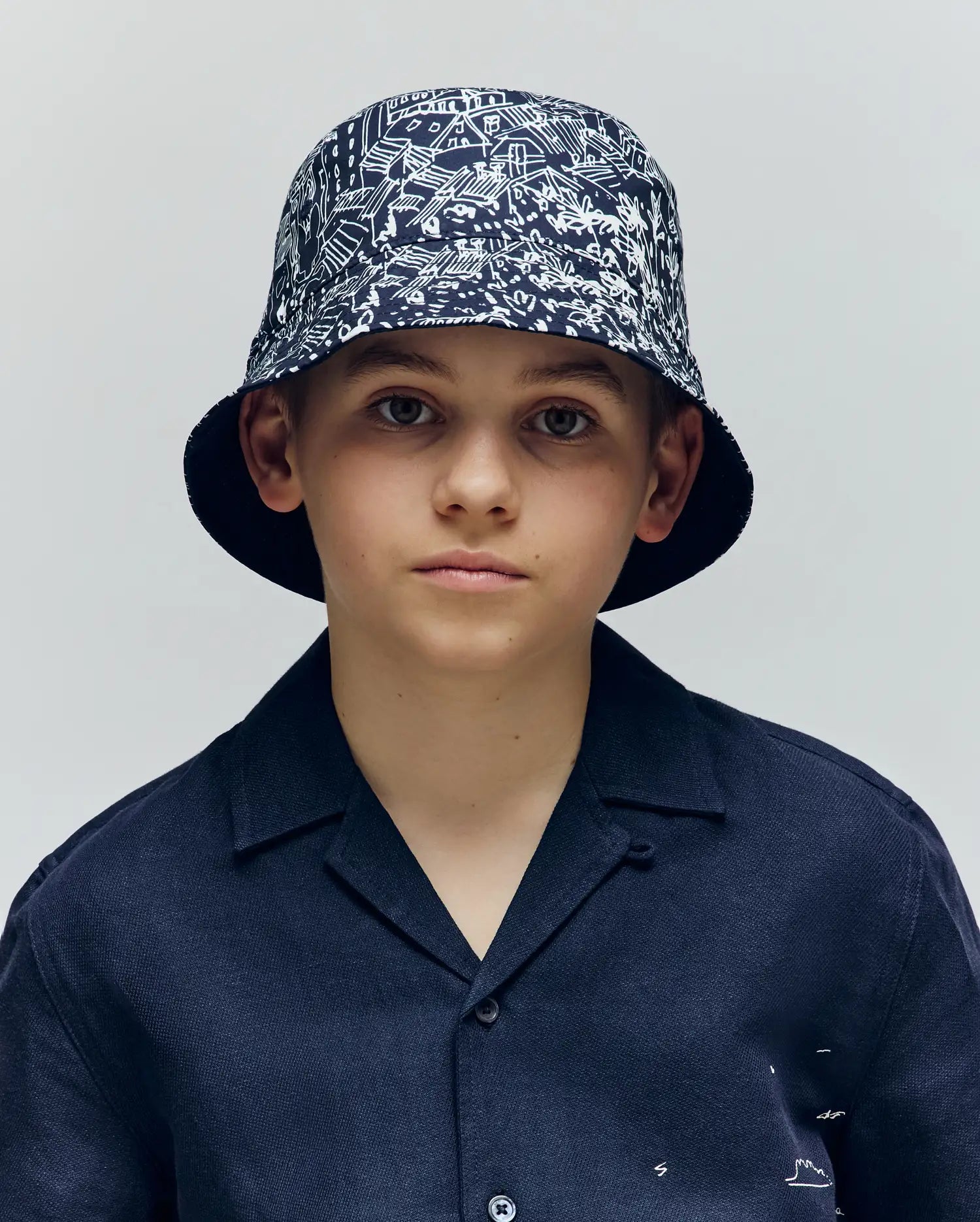 A boy wears the Psycho Bunny KIDS NAXOS REVERSIBLE BUCKET HAT - KO1500001 with black-and-white patterns and a dark short-sleeved button-up shirt, looking at the camera against a plain light background.