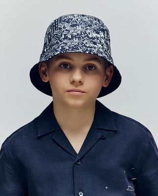 A boy wears the Psycho Bunny KIDS NAXOS REVERSIBLE BUCKET HAT - KO1500001 with black-and-white patterns and a dark short-sleeved button-up shirt, looking at the camera against a plain light background.
