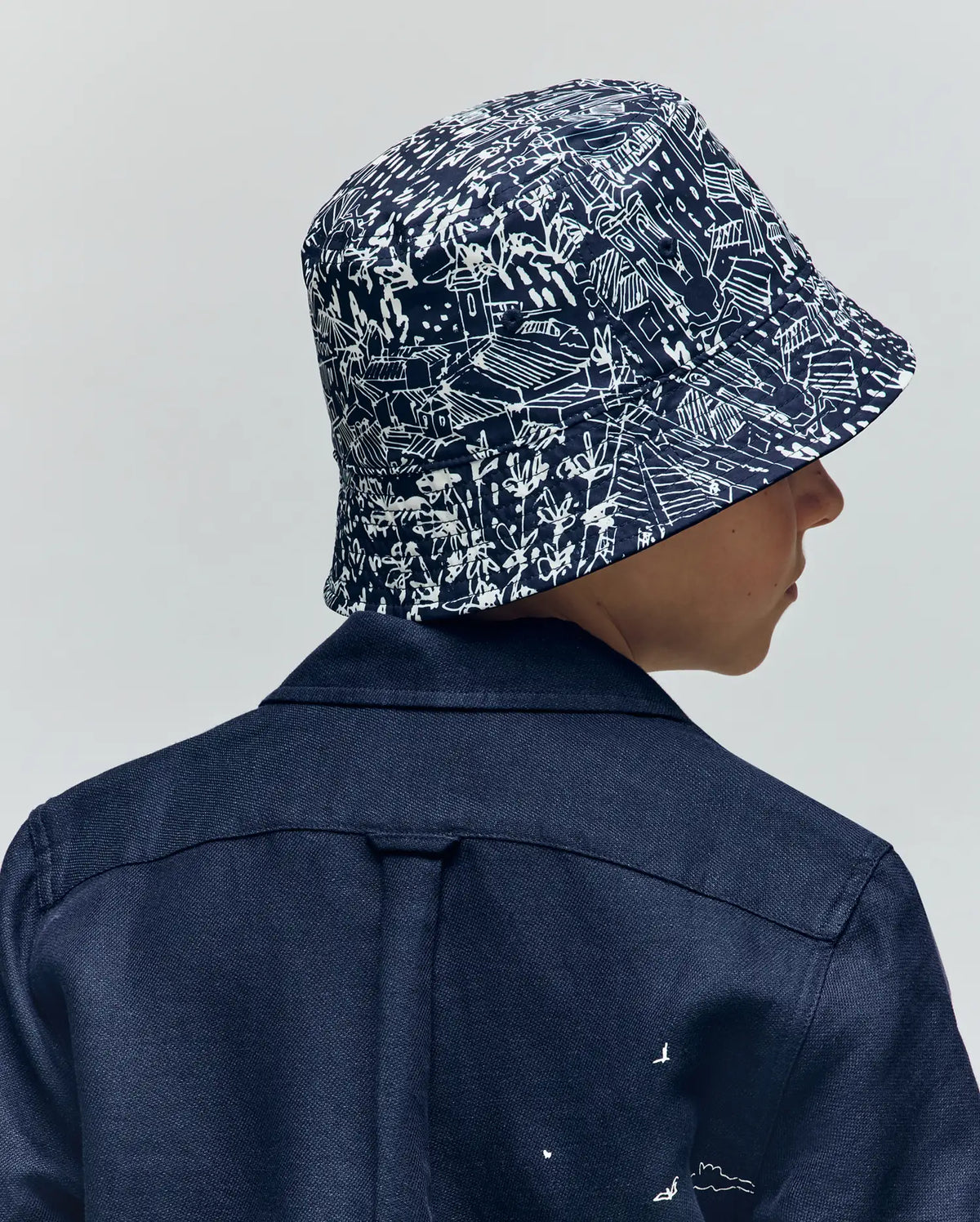 A child is seen from behind wearing a dark blue jacket and the Psycho Bunny KIDS NAXOS REVERSIBLE BUCKET HAT (KO1500001), featuring a white abstract pattern, set against a plain light background.