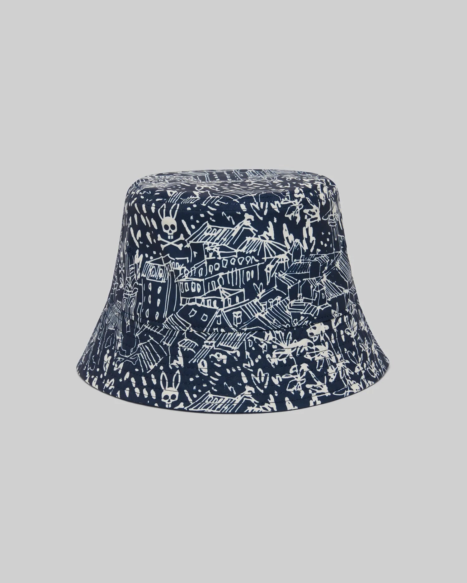 The Psycho Bunny KIDS NAXOS REVERSIBLE BUCKET HAT - KO1500001 features a navy side with white hand-drawn cityscape, buildings, rooftops, skulls, bunnies, and embroidered vents; reverse is a plain gray.