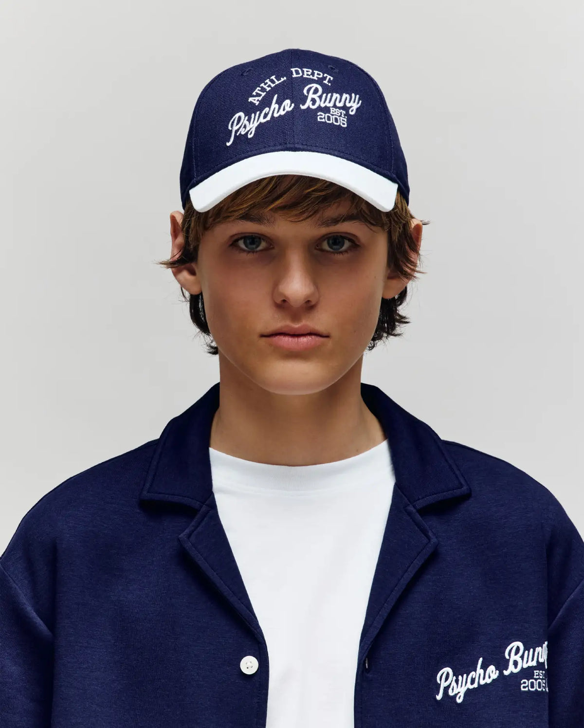 A young person sports a Psycho Bunny KIDS MATTEO BASEBALL CAP (KO1500002) in navy cotton twill with an adjustable strap, teamed with a matching jacket over a white t-shirt. Both the cap and jacket feature bold Psycho Bunny branding.