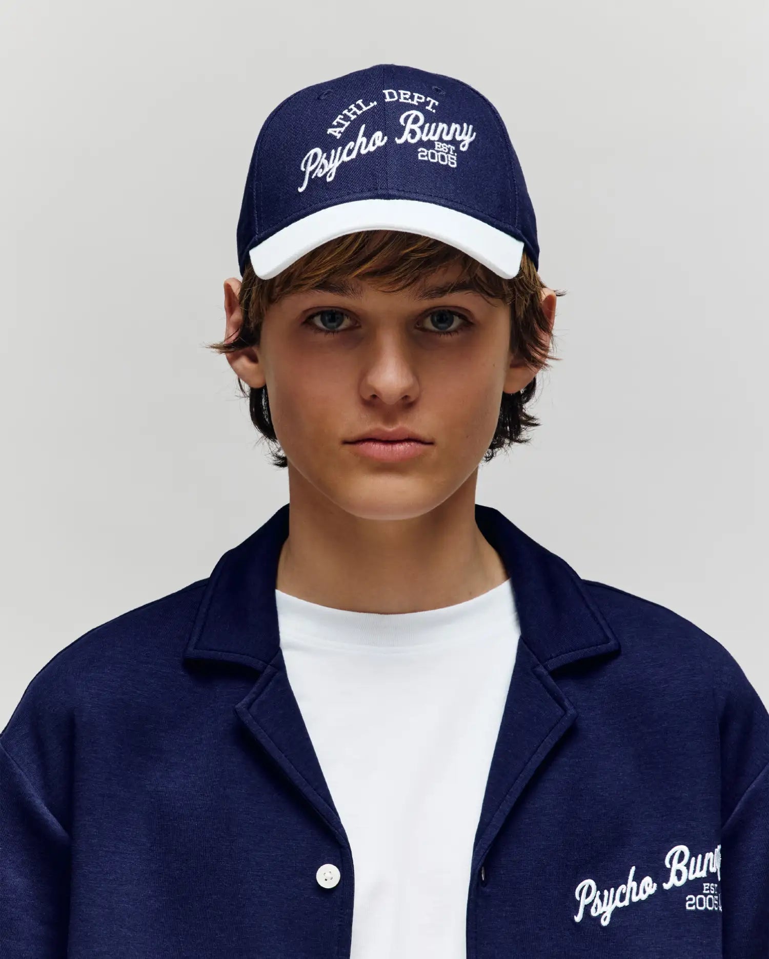 A young person sports a Psycho Bunny KIDS MATTEO BASEBALL CAP (KO1500002) in navy cotton twill with an adjustable strap, teamed with a matching jacket over a white t-shirt. Both the cap and jacket feature bold Psycho Bunny branding.
