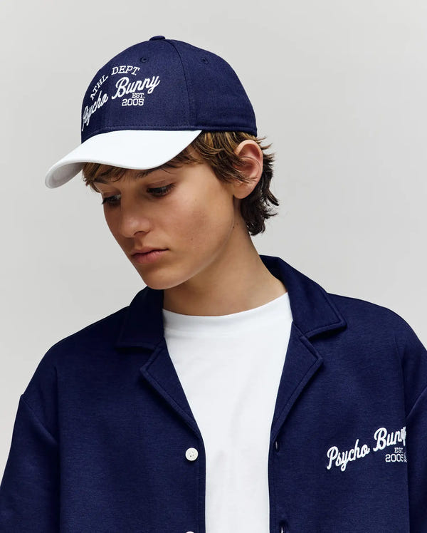 A young person wears Psycho Bunny's KIDS MATTEO BASEBALL CAP (KO1500002), a navy and white cotton twill cap with bold varsity-style embroidery and an adjustable strap, gazing down with a neutral expression against a plain backdrop.