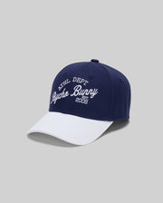 The KIDS MATTEO BASEBALL CAP by Psycho Bunny (KO1500002) is a navy blue cotton twill cap with a white brim, bold varsity-style "ATHL DEPT Psycho Bunny EST. 2005" embroidery, and an adjustable strap, shown on a light gray background.