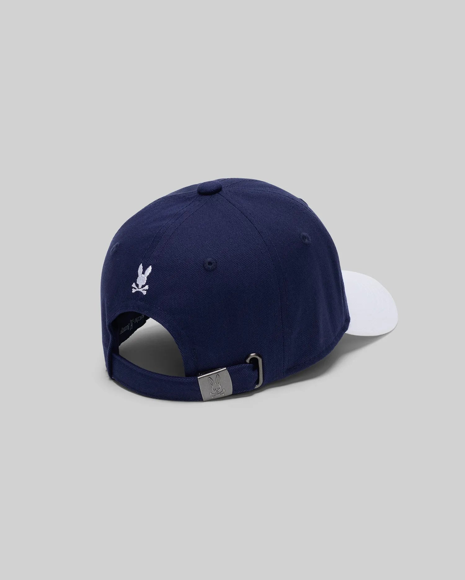The Psycho Bunny KIDS MATTEO BASEBALL CAP (KO1500002) features a varsity-style navy blue cotton twill with a white brim, small white embroidered logo above the adjustable metal buckle strap, shown from the back on a light gray background.