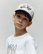 A young boy with curly brown hair wears the Psycho Bunny KIDS MATTEO BASEBALL CAP (KO1500002), a cream and navy cotton twill varsity-style hat, and a white bunny graphic shirt, standing against a plain light background.