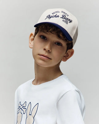 A young boy with curly brown hair wears the Psycho Bunny KIDS MATTEO BASEBALL CAP (KO1500002), a cream and navy cotton twill varsity-style hat, and a white bunny graphic shirt, standing against a plain light background.