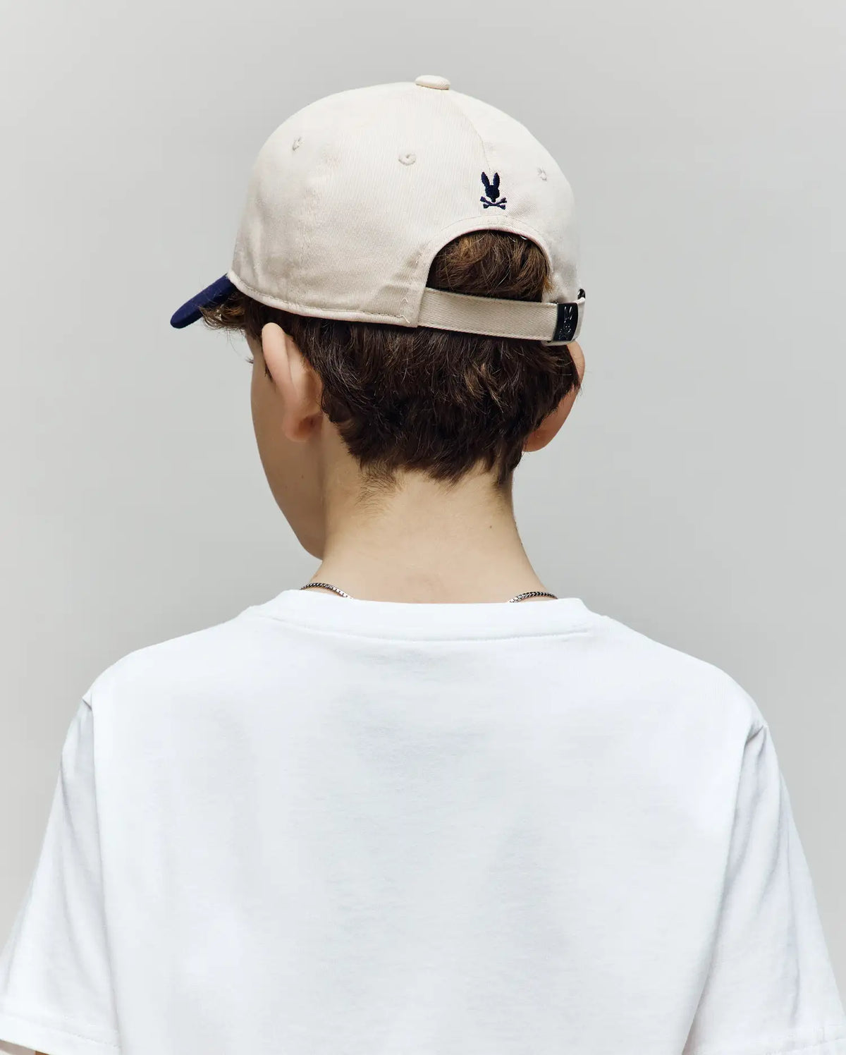 A child with short brown hair, in a white t-shirt and the Psycho Bunny KIDS MATTEO BASEBALL CAP (KO1500002) in beige with a small black logo, is shown from behind against a plain light gray background.