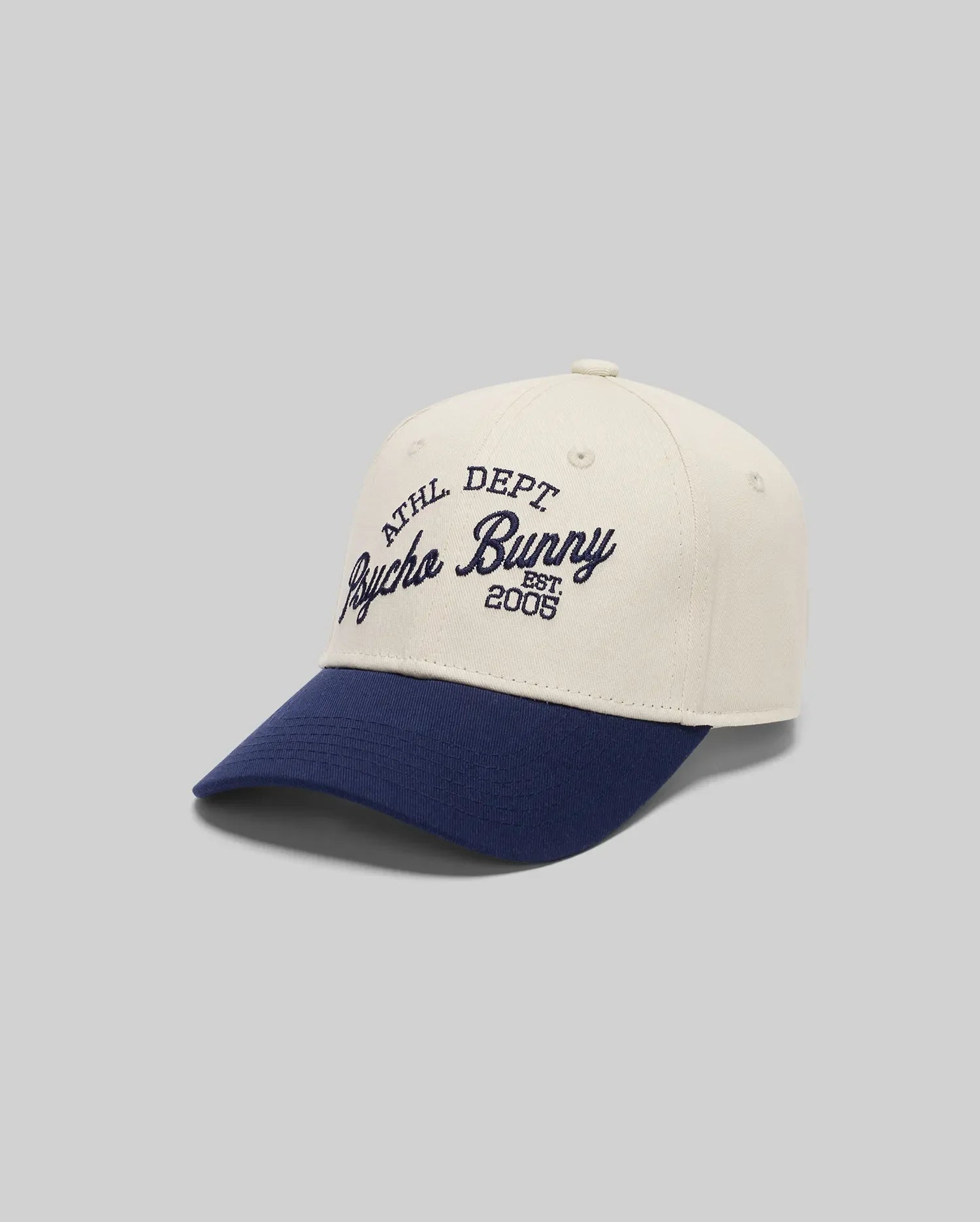 The KIDS MATTEO BASEBALL CAP (KO1500002) by Psycho Bunny features white cotton twill, a navy brim, varsity-style "ATHL DEPT Psycho Bunny EST. 2005" embroidery in navy, an adjustable strap, and is shown on a plain gray background.