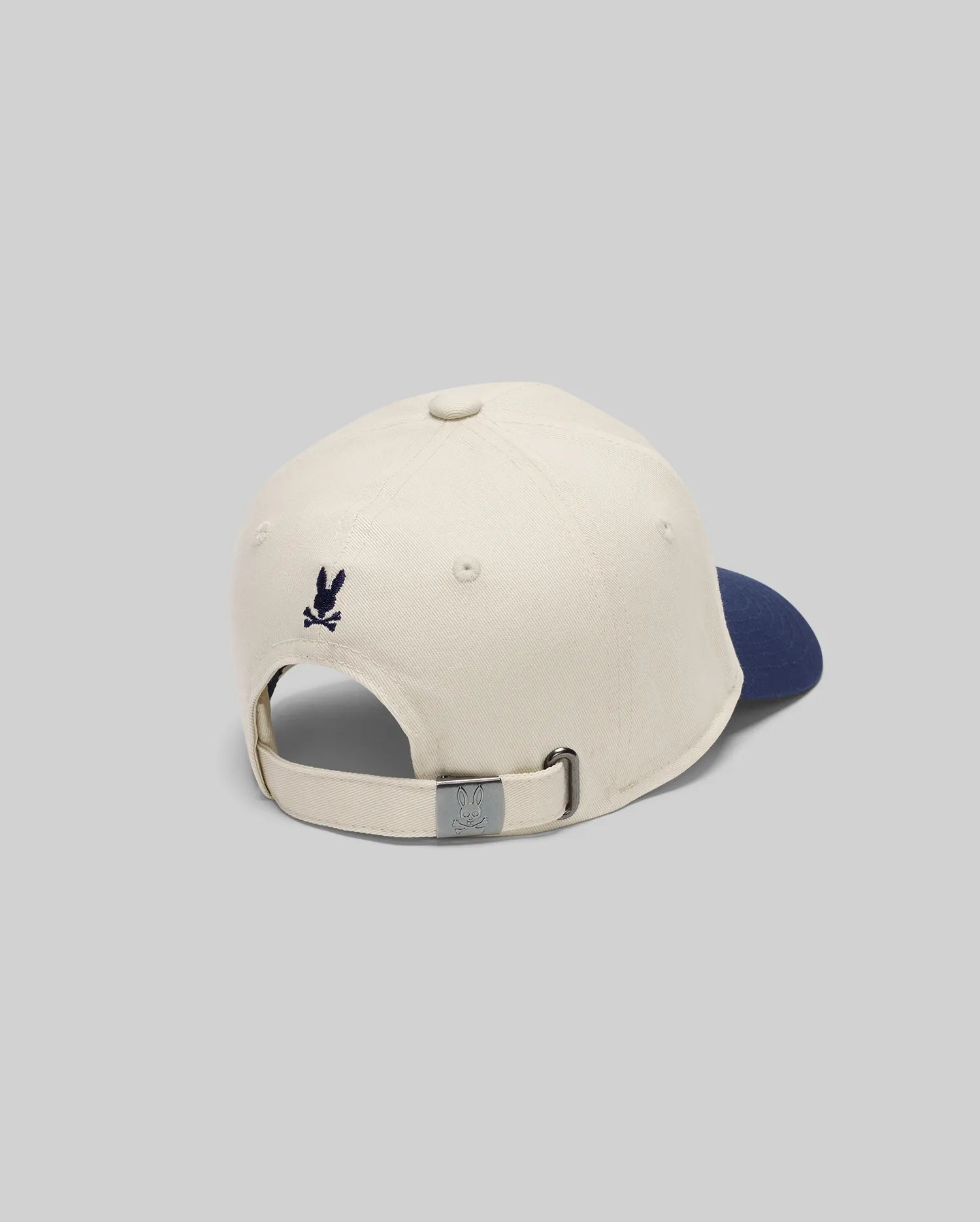 The KIDS MATTEO BASEBALL CAP (KO1500002) by Psycho Bunny is a cream cotton twill cap with a navy brim, seen from the back. It has an adjustable strap with a metal clasp and a small dark blue embroidered Psycho Bunny logo above the opening.