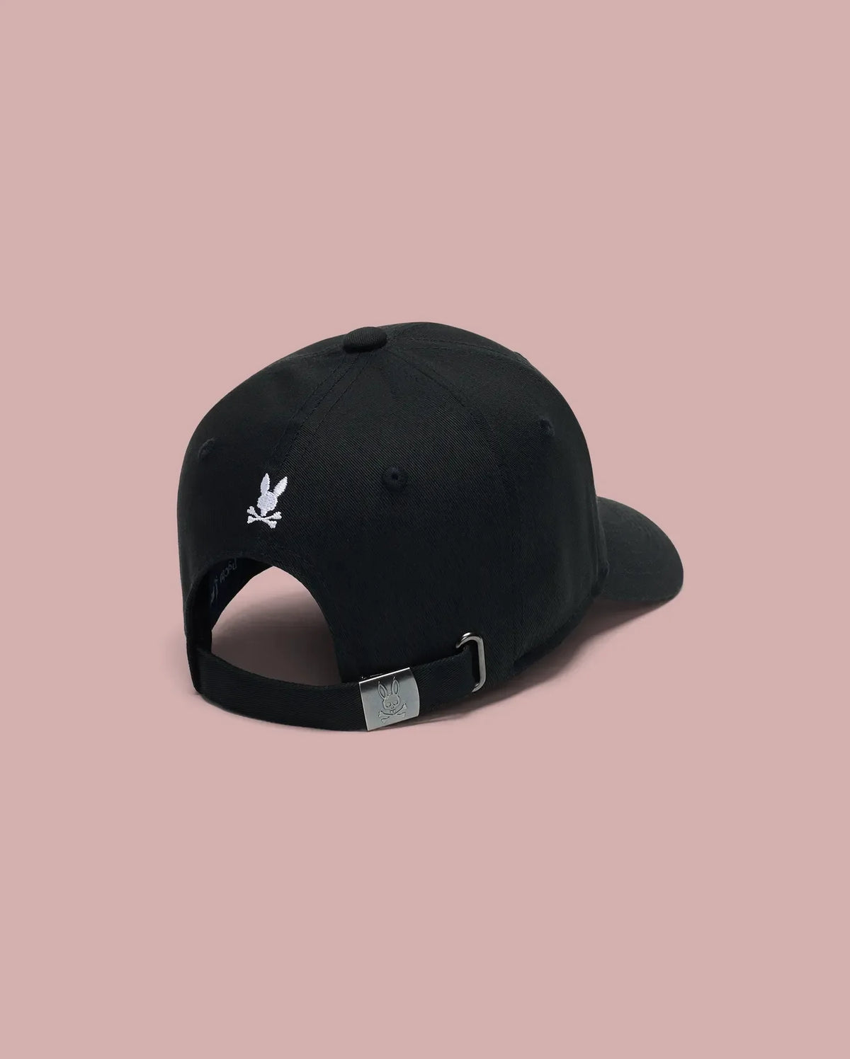 The Psycho Bunny KIDS ARCHIVE BASEBALL CAP (KO1500004) is a black cotton twill cap shown from the back, featuring a small embroidered bunny logo above the adjustable strap with an engraved silver buckle. Background is solid light pink.