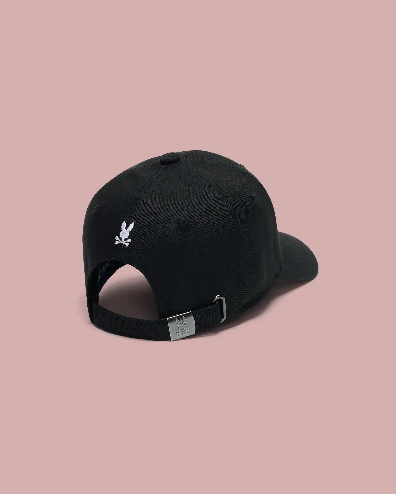 The Psycho Bunny KIDS ARCHIVE BASEBALL CAP (KO1500004) is a black cotton twill cap shown from the back, featuring a small embroidered bunny logo above the adjustable strap with an engraved silver buckle. Background is solid light pink.