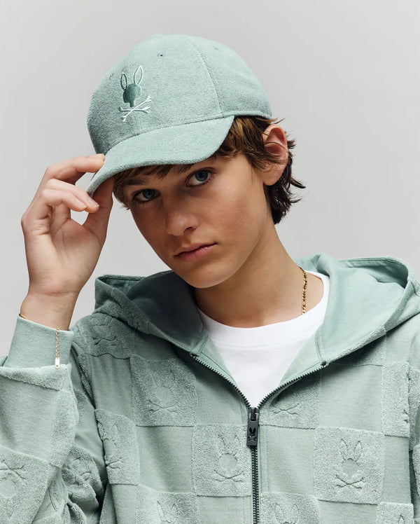 A young person in a Psycho Bunny KIDS KELVIN TOWEL TERRY BASEBALL CAP - KO1500007 and matching zip-up hoodie, featuring an embroidered Bunny logo, looks at the camera while slightly tipping the cap against a plain background.