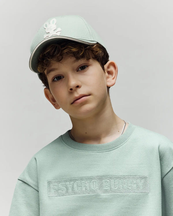 A boy in a light green Psycho Bunny premium cotton sweatshirt and a matching KIDS DEREK BASEBALL CAP (KO1500008) with the brand's bunny logo stands calmly against a gray background, looking at the camera.