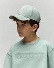 A young boy wears a light green Psycho Bunny cotton sweatshirt and the KIDS DEREK BASEBALL CAP (KO1500008) featuring a white bunny logo, looking to the side against a plain background.