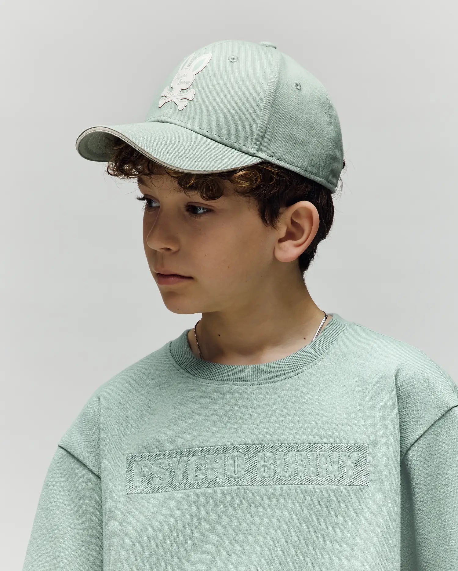 A young boy wears a light green Psycho Bunny cotton sweatshirt and the KIDS DEREK BASEBALL CAP (KO1500008) featuring a white bunny logo, looking to the side against a plain background.