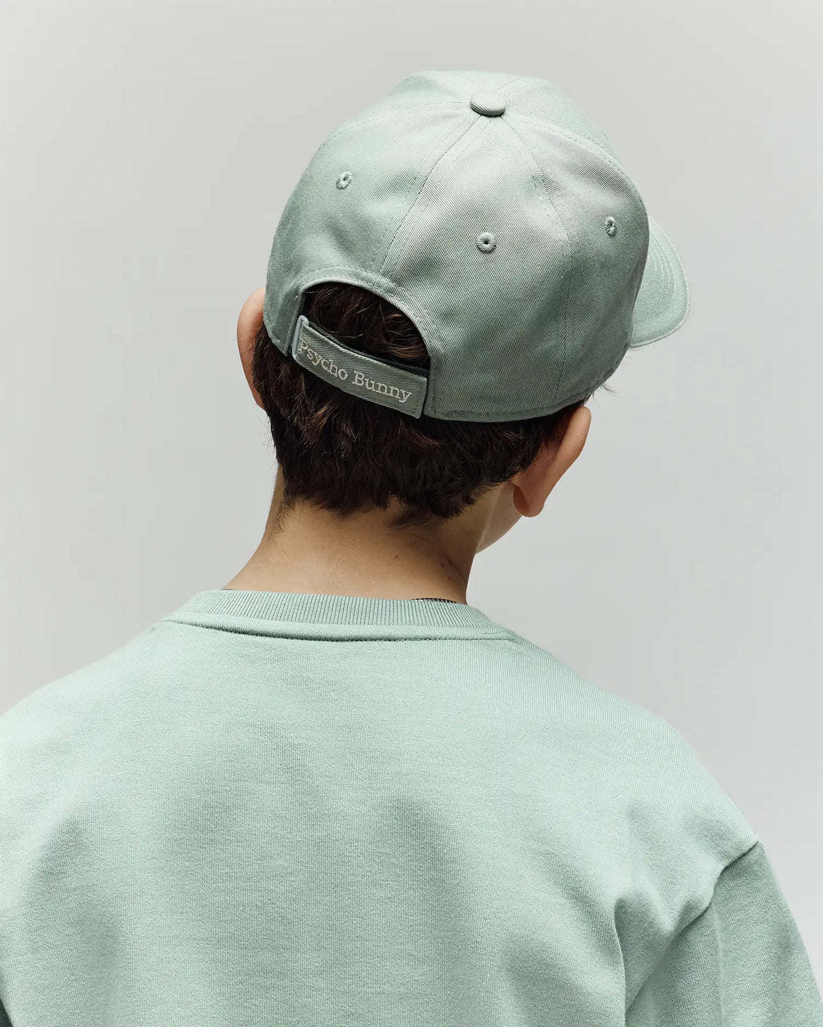 A child is seen from behind wearing a light green Psycho Bunny KIDS DEREK BASEBALL CAP (KO1500008) and a matching premium cotton sweatshirt. The cap features a "Psycho Bunny" label on its adjustable strap, set against a plain, light-colored background.