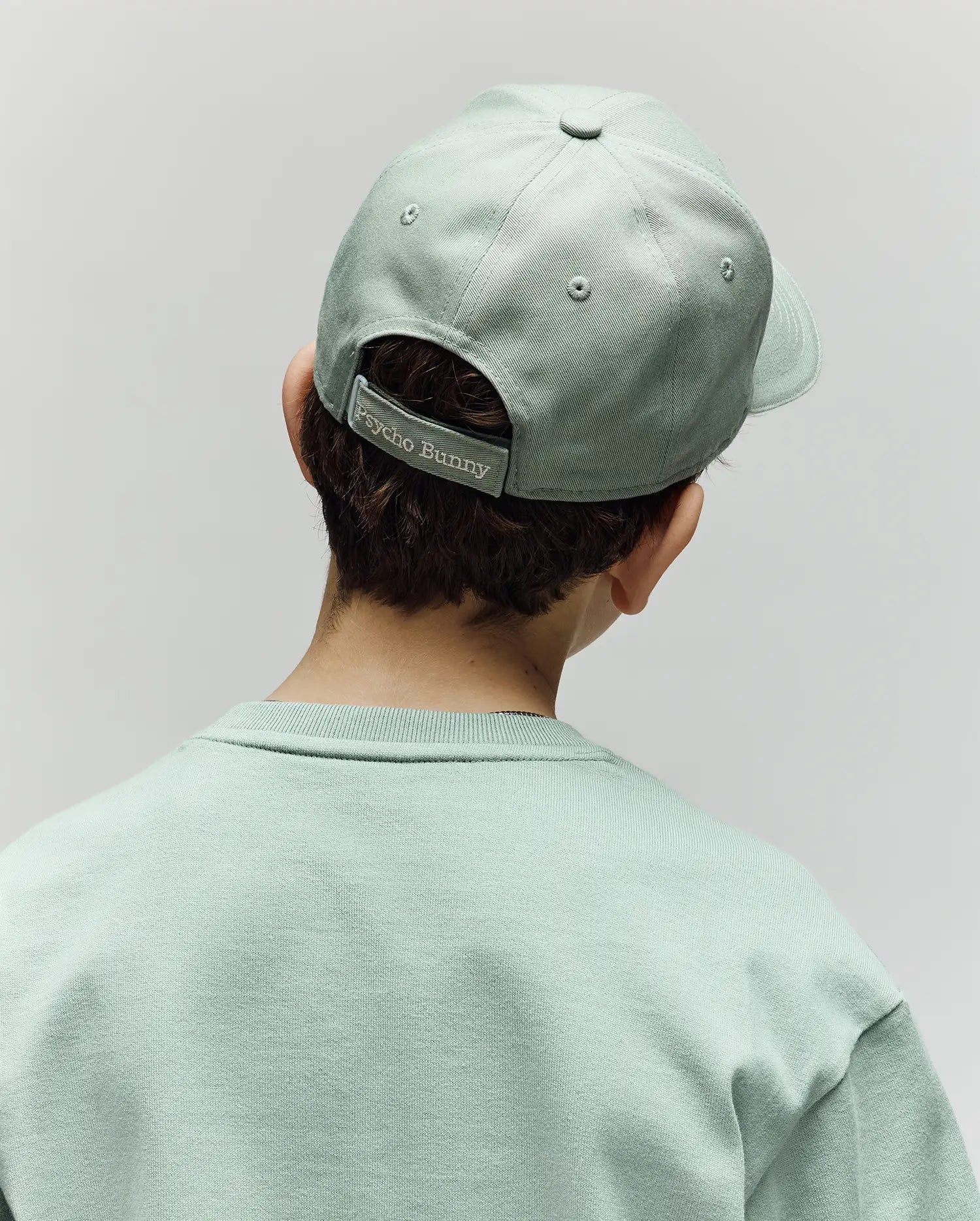 A child is seen from behind wearing a light green Psycho Bunny KIDS DEREK BASEBALL CAP (KO1500008) and a matching premium cotton sweatshirt. The cap features a "Psycho Bunny" label on its adjustable strap, set against a plain, light-colored background.
