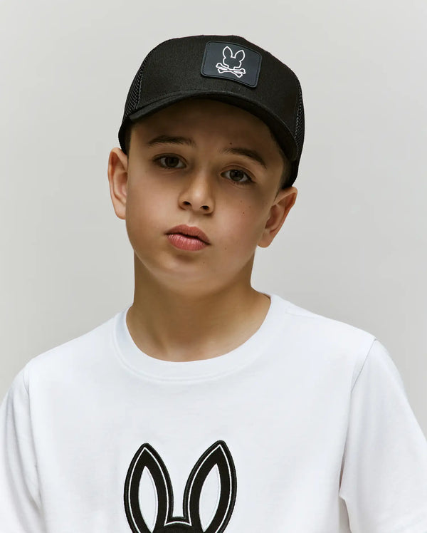 A boy wearing a Psycho Bunny KIDS DARIO TRUCKER CAP (KO1500009), a black snap-back cap with a bunny logo, and a white t-shirt with an outlined bunny graphic stands against a light background, looking at the camera.