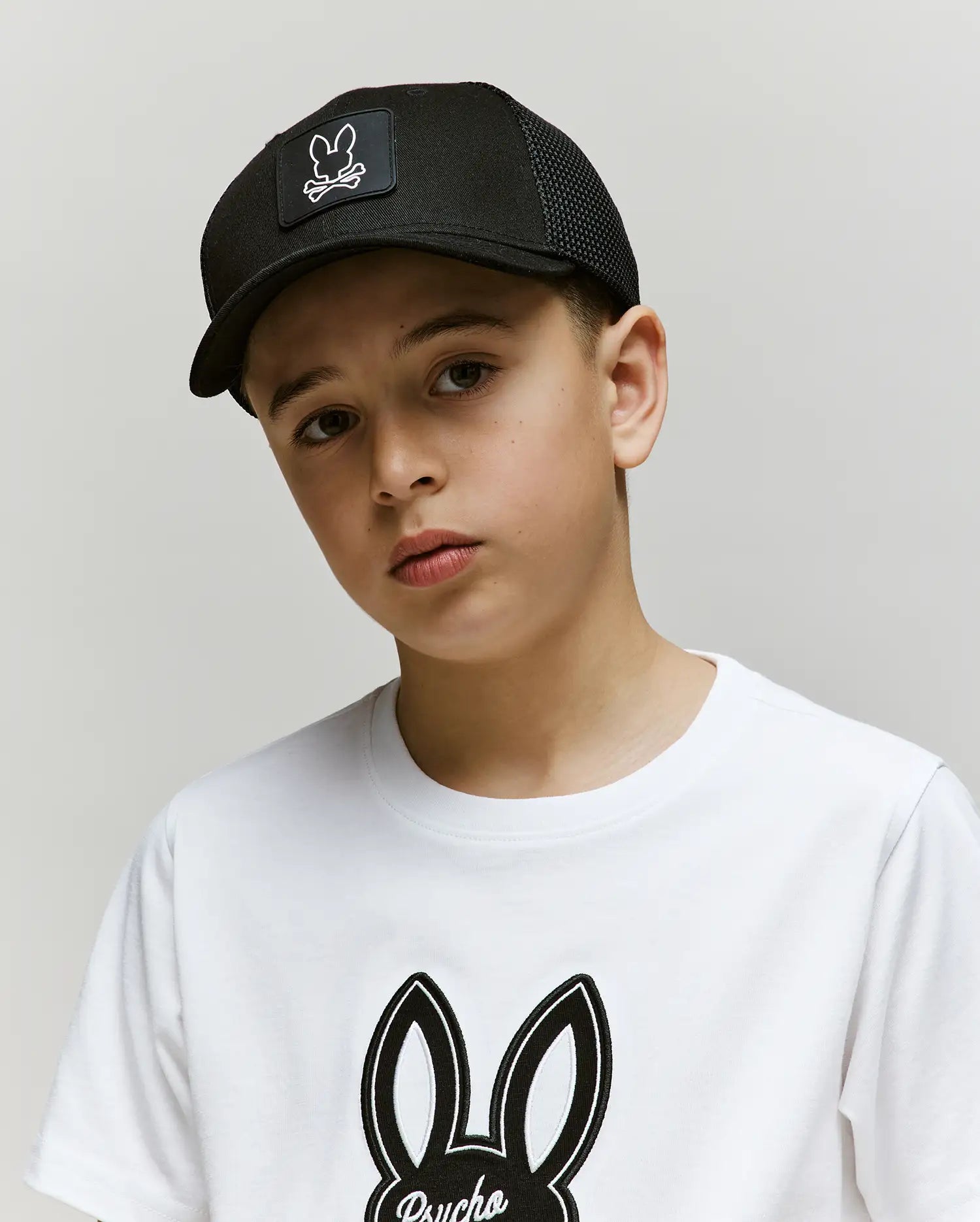 A young boy wears the Psycho Bunny KIDS DARIO TRUCKER CAP (KO1500009), a breathable black baseball cap, and a white Pima cotton T-shirt with a black bunny logo, standing against a plain light background and looking slightly serious.