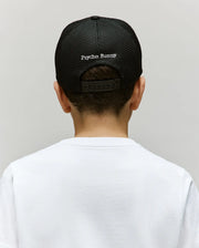A child is seen from behind in a white shirt and a black KIDS DARIO TRUCKER CAP - KO1500009 by Psycho Bunny, featuring "Psycho Bunny" embroidered in white on the adjustable snap-back, set against a plain light background.