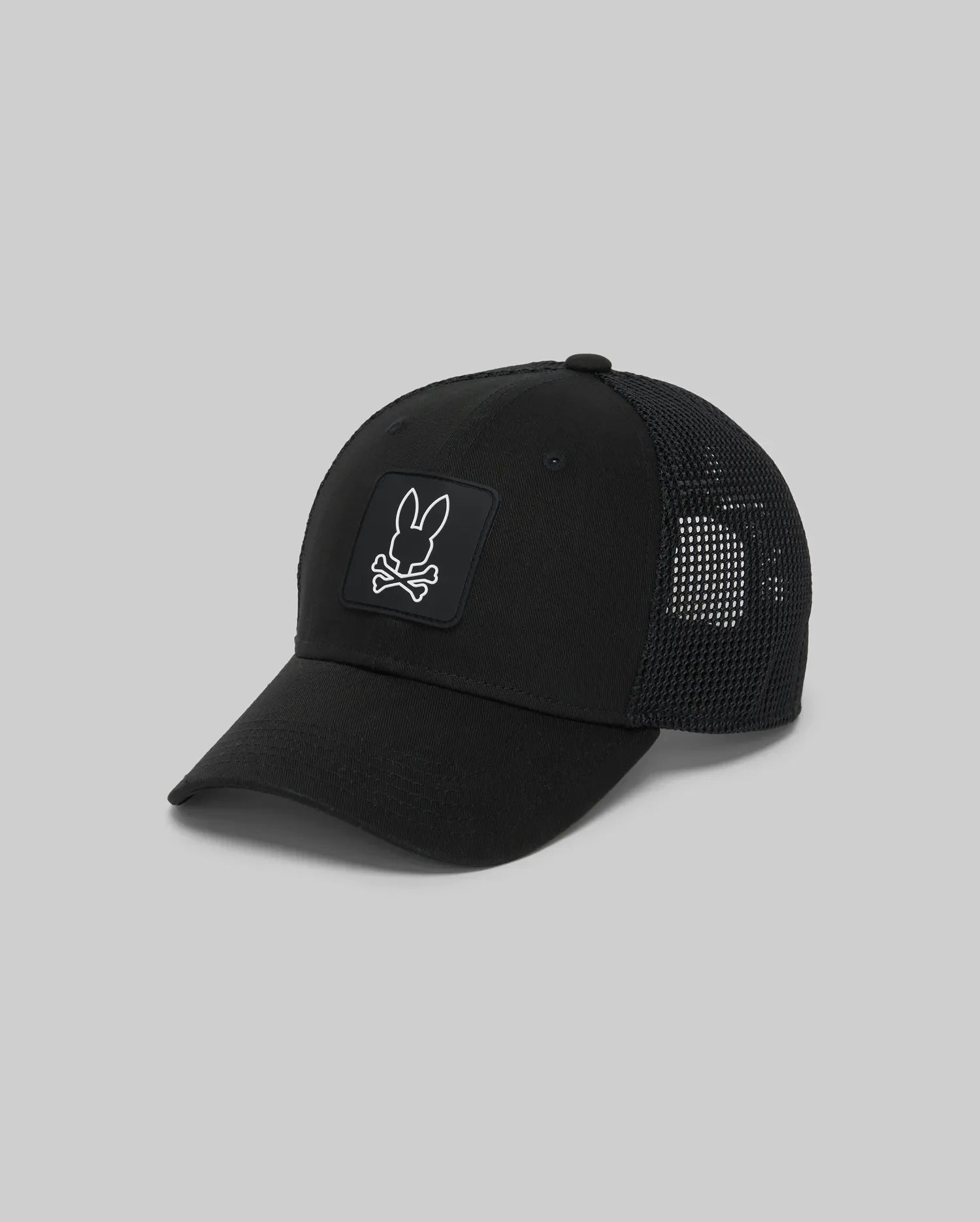 The Psycho Bunny KIDS DARIO TRUCKER CAP (KO1500009) is a black mesh hat with a curved brim, adjustable snap-back, and features a front patch of a white rabbit head above crossed bones on a black square background.