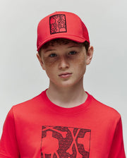 A boy with curly red hair and freckles wears a Psycho Bunny KIDS GLENN TRUCKER CAP (KO1500012) and a red T-shirt with black abstract graphics, standing against a plain light background.