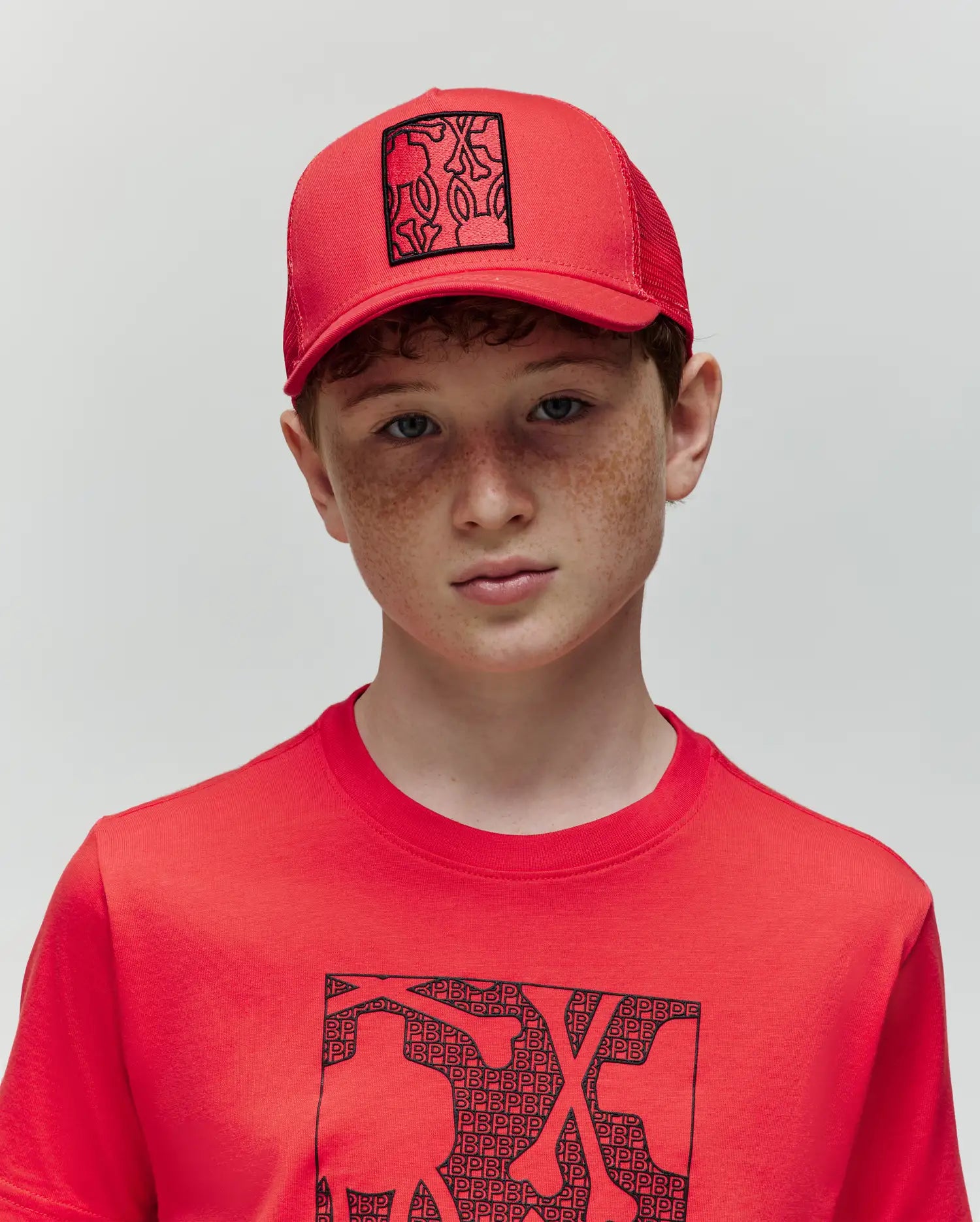 A boy with curly red hair and freckles wears a Psycho Bunny KIDS GLENN TRUCKER CAP (KO1500012) and a red T-shirt with black abstract graphics, standing against a plain light background.