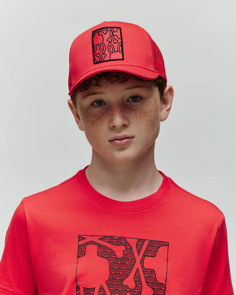 A boy with curly red hair and freckles wears a Psycho Bunny KIDS GLENN TRUCKER CAP (KO1500012) and a red T-shirt with black abstract graphics, standing against a plain light background.