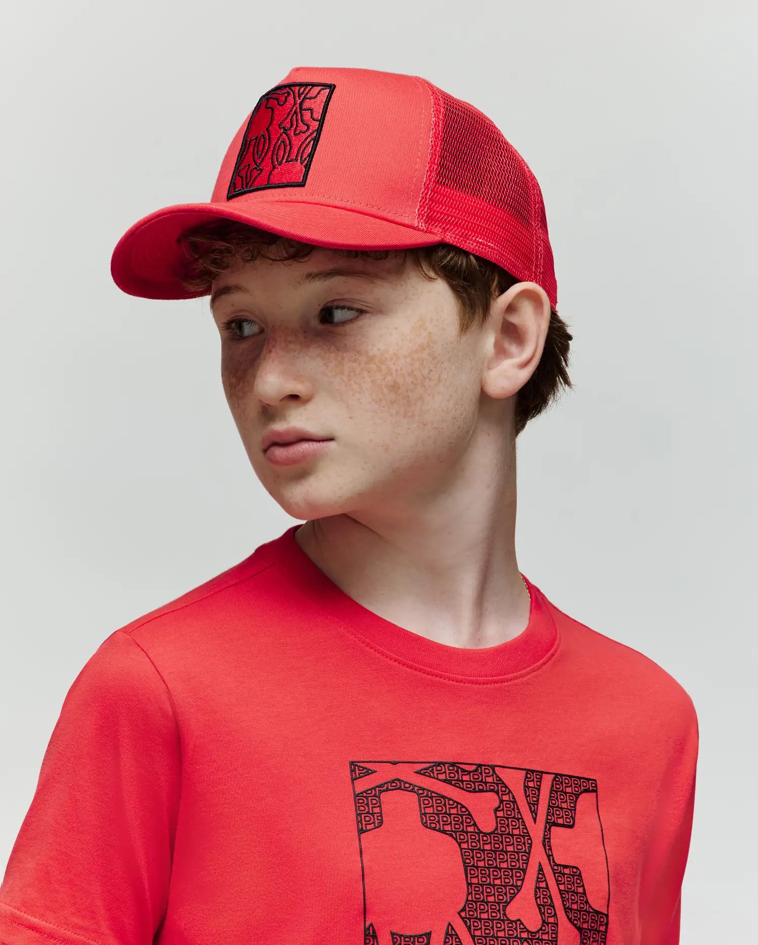 A young boy with light skin and freckles wears the Psycho Bunny KIDS GLENN TRUCKER CAP (KO1500012) and a matching red T-shirt with a black graphic, looking to his left against a plain, light background.