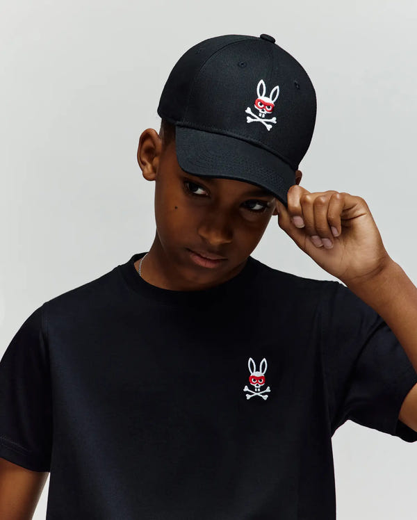 A boy in a 100% cotton black tee and the Psycho Bunny KIDS MISCHIEF BASEBALL CAP (KO1500013), both with embroidered Mischief Bunny skull-and-crossbones featuring red eyes. He tilts his cap's brim with his right hand, looking downward.